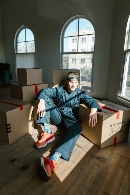 A person wearing a dark green T-shirt and dark trousers is in the process of preparing a cardboard box for a home relocation. The individual is holding the box steady with one hand while the other han