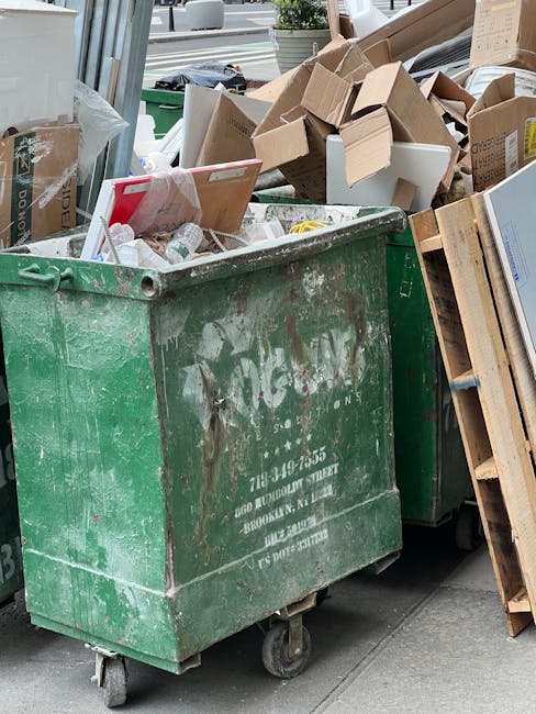 A white removal van parked on a city street next to a large, multi-storey building with a grey facade. The van's rear doors are open, revealing a load of bulky waste items inside, including black and