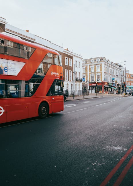 A red double-decker bus is passing along a street lined with a row of buildings, including shops and cafes, in an urban area. In the foreground, the edge of a residential driveway or loading zone is v