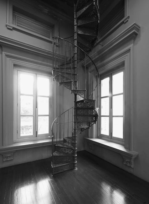 A vertically oriented view of a modern, spiral staircase inside a multi-storey building, featuring concrete steps with wooden treads and black metal railings that follow the curve of the staircase. Th