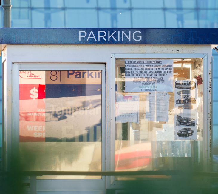 A close-up view of a public parking sign and notice board located at a bus stop or parking shelter in an urban setting. The sign at the top displays the word 'PARKING' in bold white letters on a blue